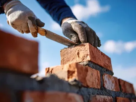 Mason in gloves laying brick on mortar for an outdoor wall under a blue sky